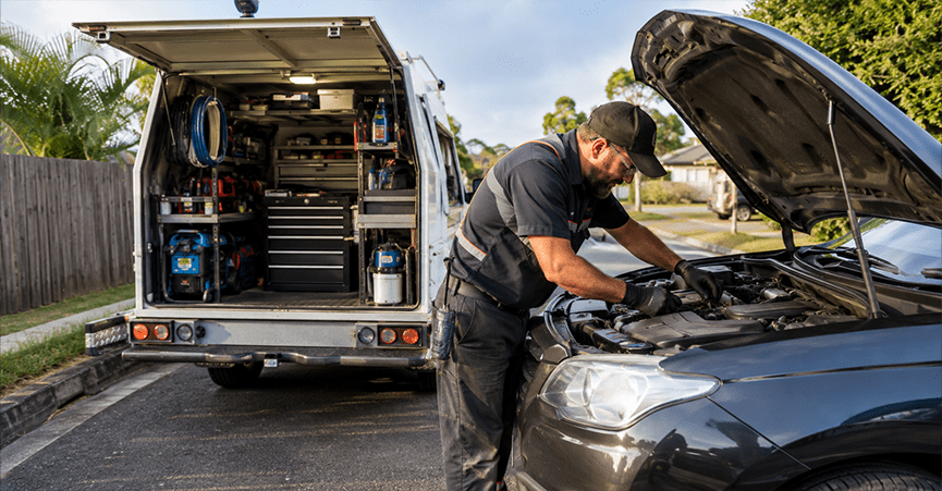 A mobile mechanic wearing a cap and gloves works on the open engine bay of a black car parked on a suburban street. Behind him, the back of his white service van is open, revealing organized shelves filled with tools and equipment.