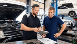 An auto mechanic shows information on a digital tablet to a customer inside a busy repair shop, with an open car hood in the foreground and other vehicles on lifts in the background.