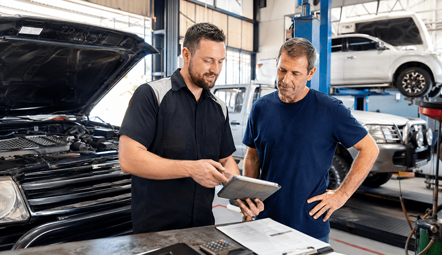 An auto mechanic shows information on a digital tablet to a customer inside a busy repair shop, with an open car hood in the foreground and other vehicles on lifts in the background.
