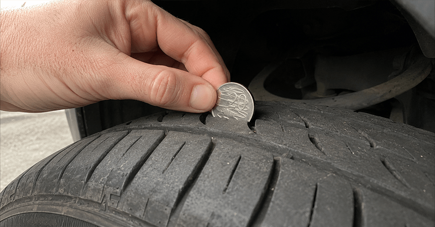 A close-up of a person's hand inserting an Australian 20-cent coin into the tread groove of a car tyre to check its depth.