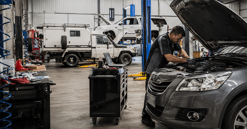 A mechanic works on the engine of a grey car with its hood open inside a large, busy auto repair shop. A black tool cart sits nearby, and other vehicles, including a white truck, are visible in the background on lifts.