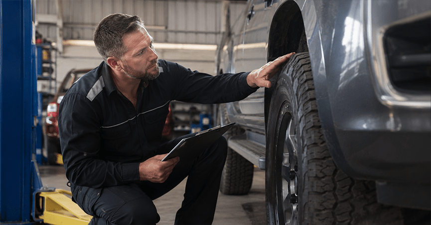 A mechanic holding a clipboard crouches down to inspect the tread of a large tyre on a grey truck inside an auto repair shop.