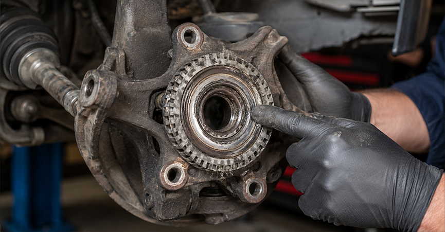 Auto mechanic pointing to a damaged wheel bearing and ABS tone ring on a car steering knuckle.